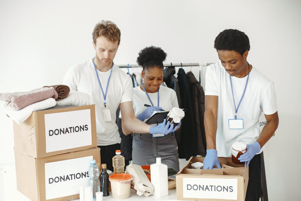 why-choose-us Three volunteers organizing donation boxes with clothes and food items in a community center.